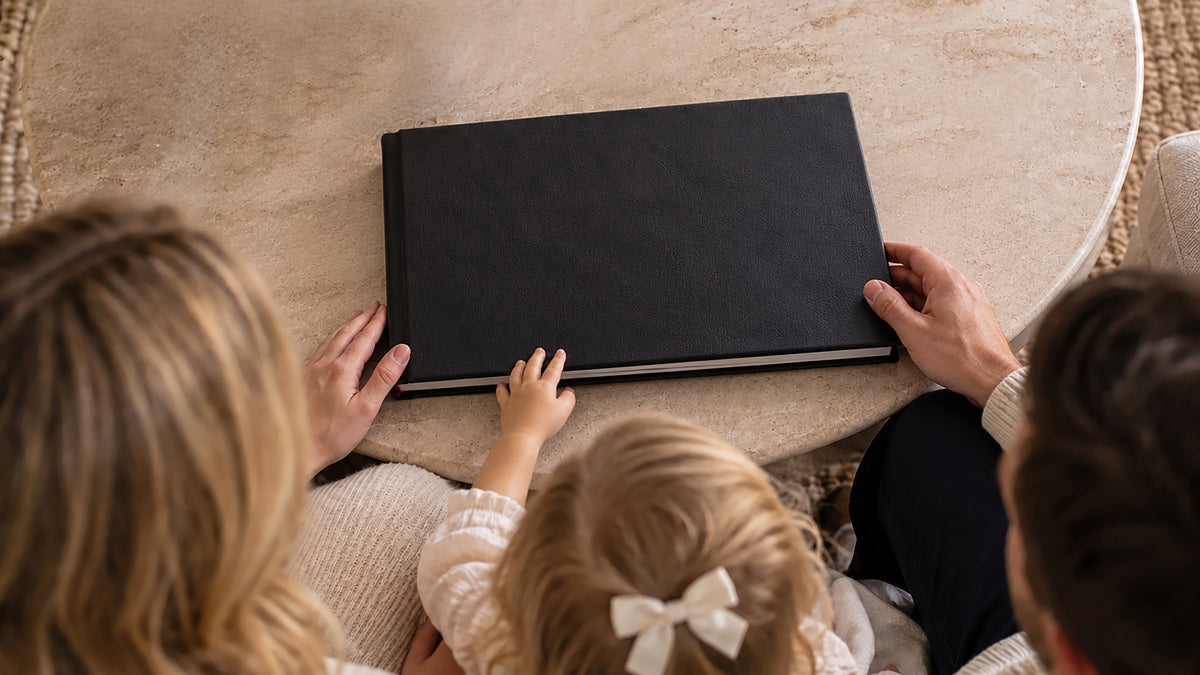 Handcrafted raven black leatherette wedding album on a coffee table, a family gathered to open it together at home